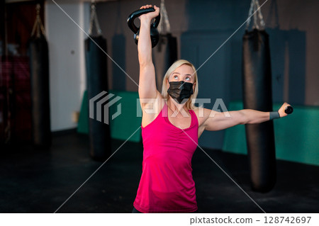 Woman lifting kettlebell overhead and extending dumbbell arm in gym, with punching bags 128742697