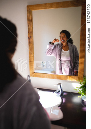 Woman brushing teeth at bathroom sink, with toothbrush, mirror, chrome faucet and soap dispenser 128742700