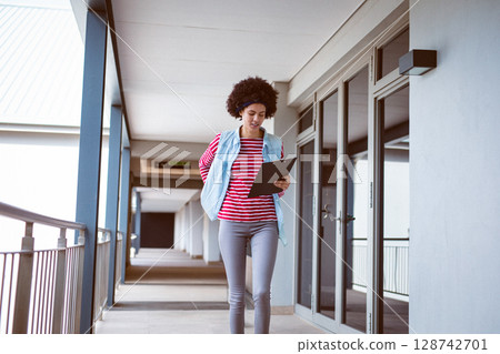 Woman walking along covered exterior corridor of modern building, holding black clipboard 128742701