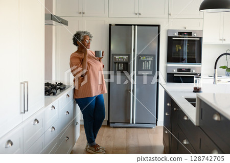 Senior woman leaning against cabinets in kitchen by refrigerator, holding ceramic mug, copy space 128742705