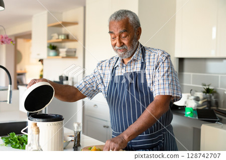 Senior man lifting compost bin lid while chopping bell peppers and herbs at modern kitchen island Senior man lifting compost bin lid while chopping bell peppers and herbs at modern kitchen island 128742707