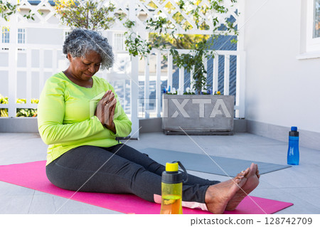 Senior woman sitting on pink yoga mat on tiled balcony, holding prayer pose with water bottle Senior woman sitting on pink yoga mat on tiled balcony, holding prayer pose with water bottle 128742709