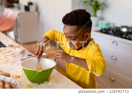 African American boy whisking eggs with metal whisk in green bowl in home kitchen, flour smudges 128742713