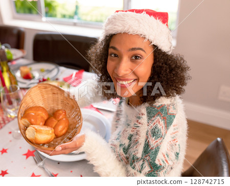 Female child wearing Santa hat sitting at dining table at home, holding wicker basket with rolls Female child wearing Santa hat sitting at dining table at home, holding wicker basket with rolls 128742715