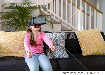 Girl sitting on dark grey couch wearing VR headset in modern living room with decorative pillows Girl sitting on dark grey couch wearing VR headset in modern living room with decorative pillows 128742719