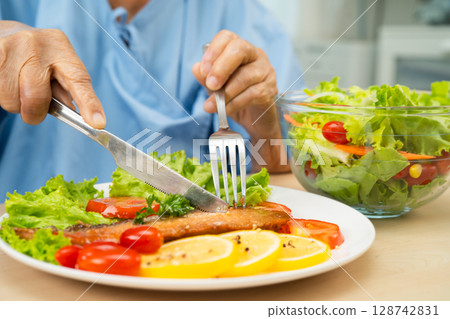 Asian elderly woman patient eating salmon stake and vegetable salad for healthy food in hospital. Asian elderly woman patient eating salmon stake and vegetable salad for healthy food in hospital. 128742831