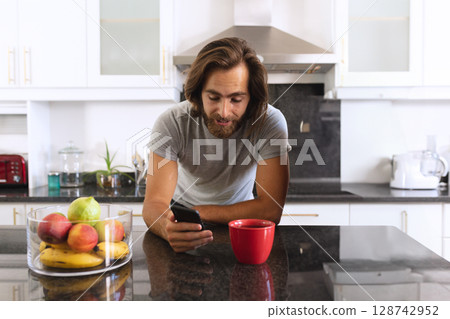 Male leaning on countertop using smartphone in kitchen, with coffee mug and glass fruit bowl 128742952