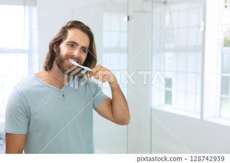 Young man brushing teeth in modern bathroom, with toothbrush and mirror, copy space 128742959