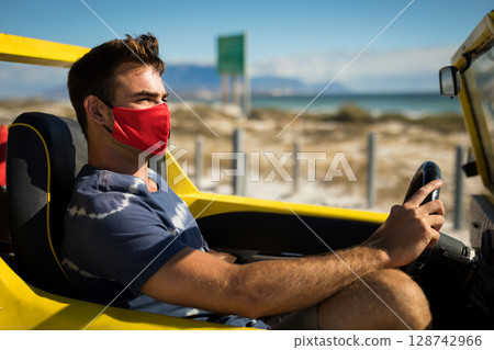 Young adult man driving yellow convertible on coastal highway with red face mask 128742966