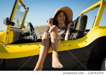 Young woman wearing floral dress sitting in yellow dune buggy on sandy terrain, adjusting straw hat 128742970