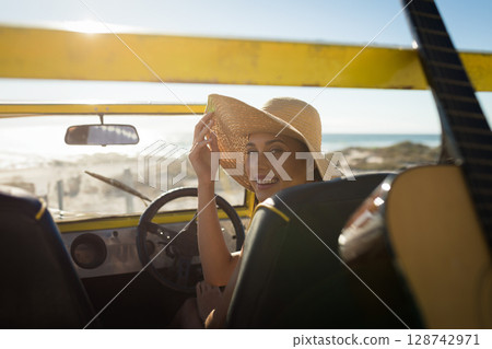 woman sitting in yellow dune buggy at sandy beach, adjusting straw hat and reaching toward guitar 128742971