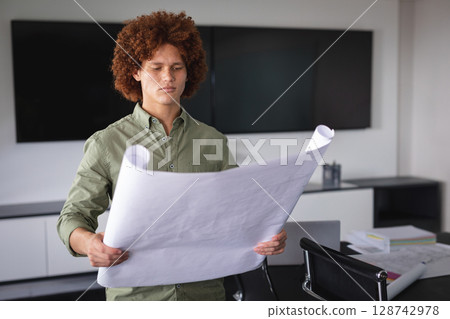 Young adult man reviewing architectural blueprint in conference room with monitors and papers Young adult man reviewing architectural blueprint in conference room with monitors and papers 128742978