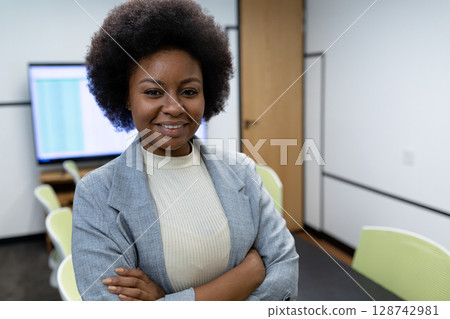 African American woman standing with arms crossed in office with green chairs, flat-screen monitor 128742981