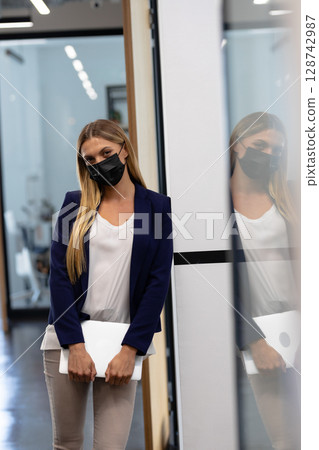Businesswoman standing in office hallway holding silver laptop with glass partition reflection Businesswoman standing in office hallway holding silver laptop with glass partition reflection 128742987
