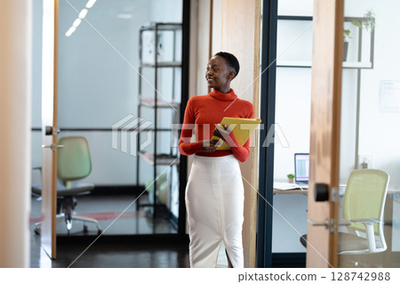 Young African American woman standing in office corridor, holding yellow folder beside open laptop 128742988