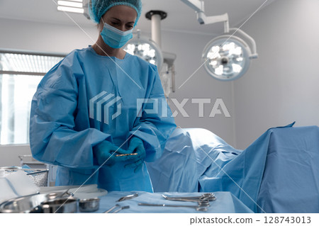 Female surgeon preparing instruments in operating room, using metal tray under surgical lights 128743013