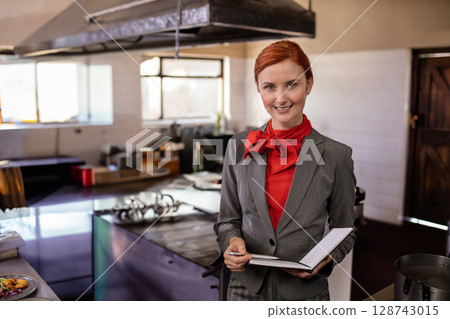 female kitchen manager holding notebook, pen in commercial kitchen with steel counters, copy space 128743015