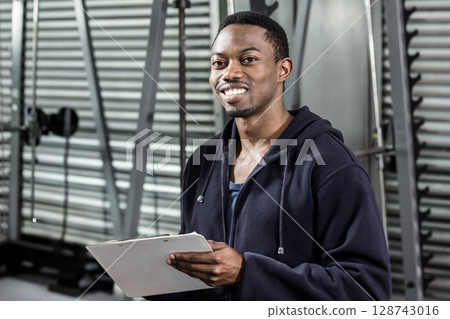 Young African American gym trainer standing in gym, with clipboard, weight machines and squat rack Young African American gym trainer standing in gym, with clipboard, weight machines and squat rack 128743016
