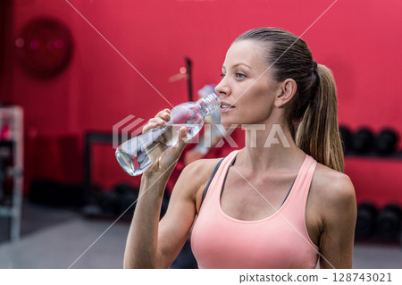 Fit woman drinking from water bottle in gym weight area with dumbbell rack, copy space 128743021