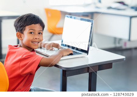 Boy typing on laptop at desk smiling at camera in classroom, with orange chairs, copy space 128743029