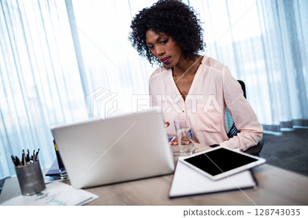 African American woman typing on laptop at office desk, with tablet, glass of water, copy space 128743035