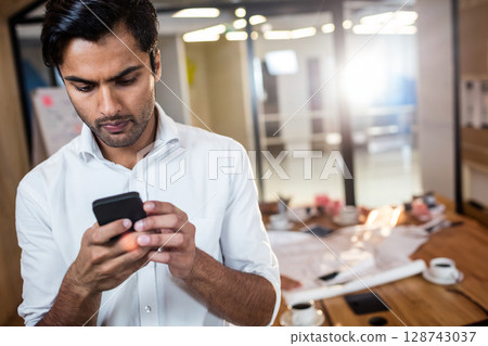 Asian man tapping smartphone in conference room by wooden table with documents and coffee cups Asian man tapping smartphone in conference room by wooden table with documents and coffee cups 128743037