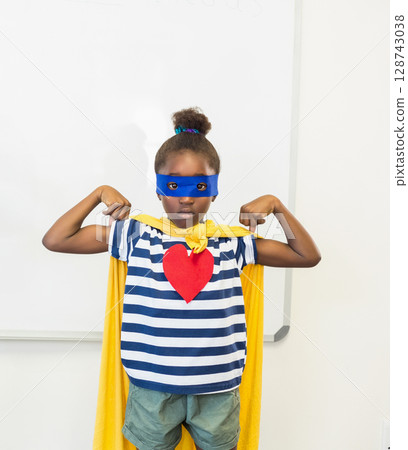 African American girl flexing arms in classroom at whiteboard, with yellow cape and blue eye mask African American girl flexing arms in classroom at whiteboard, with yellow cape and blue eye mask 128743038