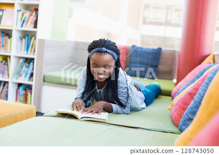African American teenage girl reading open book on cushioned bench in library nook, smiling African American teenage girl reading open book on cushioned bench in library nook, smiling 128743050
