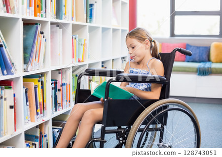 African American teenage girl sitting in manual wheelchair in library aisle reading open green book African American teenage girl sitting in manual wheelchair in library aisle reading open green book 128743051