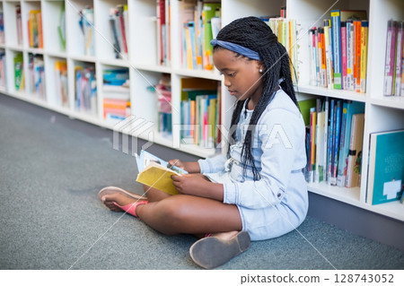 Teenage African American girl sitting on carpet reading small hardcover book in library, copy space Teenage African American girl sitting on carpet reading small hardcover book in library, copy space 128743052