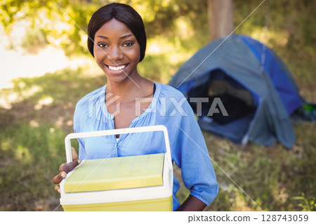 Young African American woman holding cooler near camping tent at campsite, enjoying sunny day 128743059