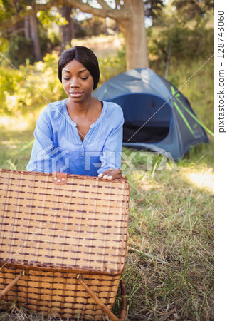 Young African American woman kneeling on campsite opening wicker picnic basket under sunlit trees Young African American woman kneeling on campsite opening wicker picnic basket under sunlit trees 128743060