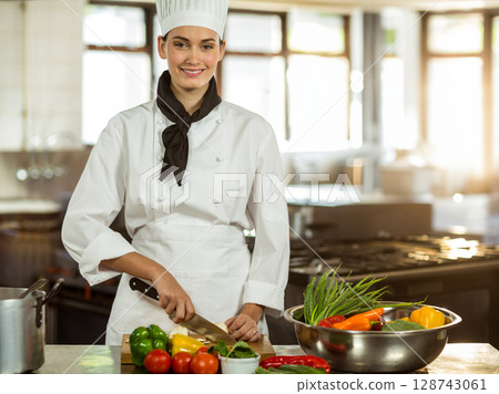 Young female chef slicing vegetables with chef's knife on wooden board in kitchen, mixing bowl Young female chef slicing vegetables with chef's knife on wooden board in kitchen, mixing bowl 128743061