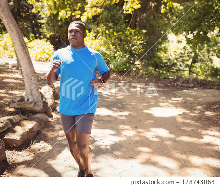 African American man jogging along sunlit trail in wooded park, with smartwatch and running shoes 128743063