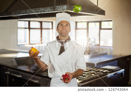 Mid adult male chef juggling colorful bell peppers in commercial kitchen, showing cooking skill Mid adult male chef juggling colorful bell peppers in commercial kitchen, showing cooking skill 128743064