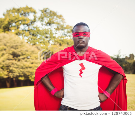 African American man standing in park wearing red cape and superhero mask with lightning-bolt logo 128743065