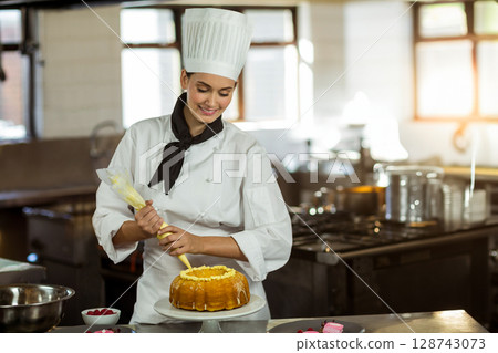 Female pastry chef piping icing onto bundt cake in commercial kitchen with pastry bag, raspberries Female pastry chef piping icing onto bundt cake in commercial kitchen with pastry bag, raspberries 128743073