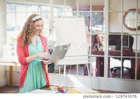 Woman standing at desk in modern office holding silver laptop and smiling by white flip chart Woman standing at desk in modern office holding silver laptop and smiling by white flip chart 128743076