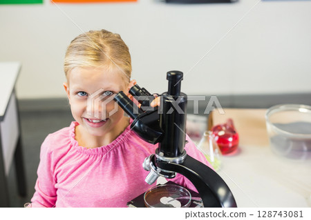 Young boy peering into black microscope at school lab bench, with petri dish, flask and bowl 128743081