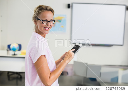 Female teacher standing in bright classroom holding tablet, with globe and world map, copy space Female teacher standing in bright classroom holding tablet, with globe and world map, copy space 128743089