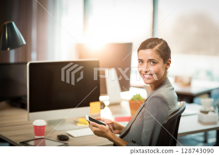 woman holding smartphone smiling at open plan office desk with monitor and coffee cup, copy space woman holding smartphone smiling at open plan office desk with monitor and coffee cup, copy space 128743090