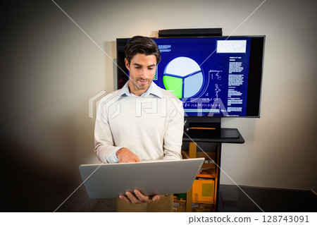 Man holding laptop and interacting with pie chart on large monitor in office showcasing data 128743091