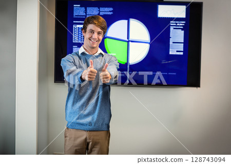 Man giving thumbs up in conference room, presenting pie chart on wall-mounted display 128743094