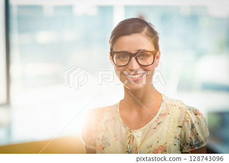 Young adult woman standing and smiling in office with black-framed glasses and floral blouse 128743096