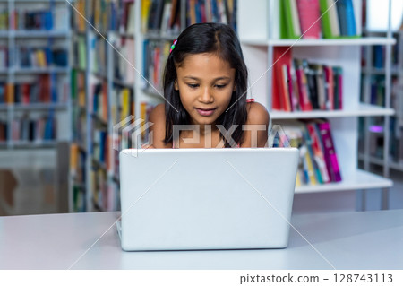 Girl child sitting at white table in library with bookshelves, working on silver laptop Girl child sitting at white table in library with bookshelves, working on silver laptop 128743113