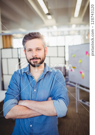 Man standing with arms crossed in open-plan office, with whiteboard holding colorful sticky notes 128743120