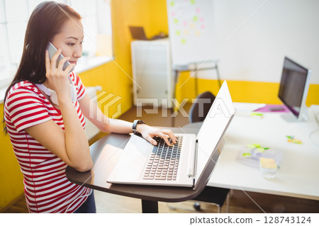 Asian woman with headphones talking on smartphone and typing on laptop in office, copy space Asian woman with headphones talking on smartphone and typing on laptop in office, copy space 128743124