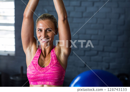 Mid adult woman raising arms overhead in gym, with blue exercise ball and weight stacks 128743133