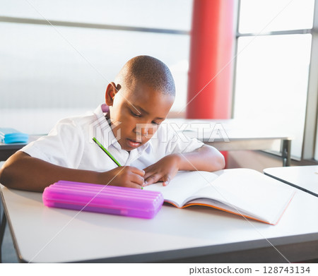 African American boy writing in notebook in classroom, using green pencil and purple pencil case African American boy writing in notebook in classroom, using green pencil and purple pencil case 128743134