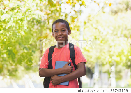Diverse male child holding blue tablet, wearing black backpack in sunlit park, smiling at camera 128743139